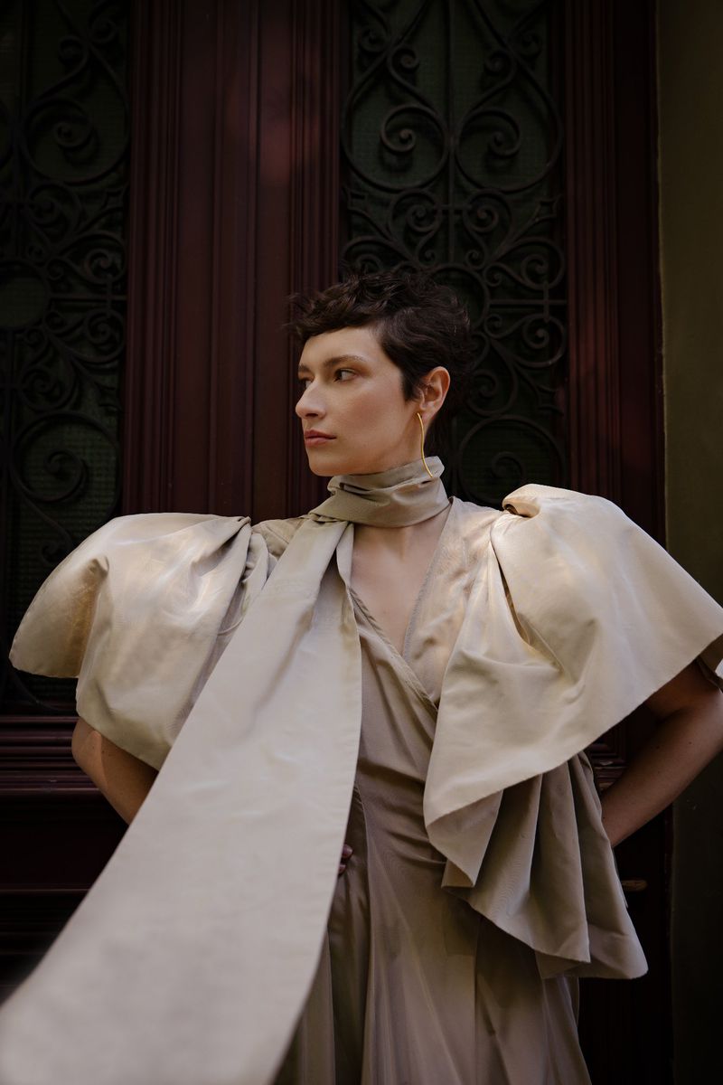 Portrait of a female model with short hair wearing an elegant beige dress with large sleeves and scarf, standing in front of a dark door.