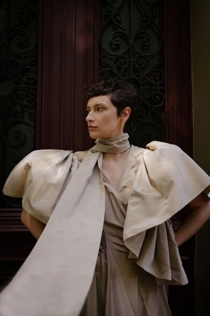 Portrait of a female model with short hair wearing an elegant beige dress with large sleeves and scarf, standing in front of a dark door.