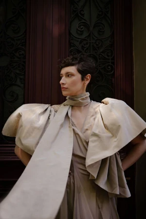 Portrait of a female model with short hair wearing an elegant beige dress with large sleeves and scarf, standing in front of a dark door.