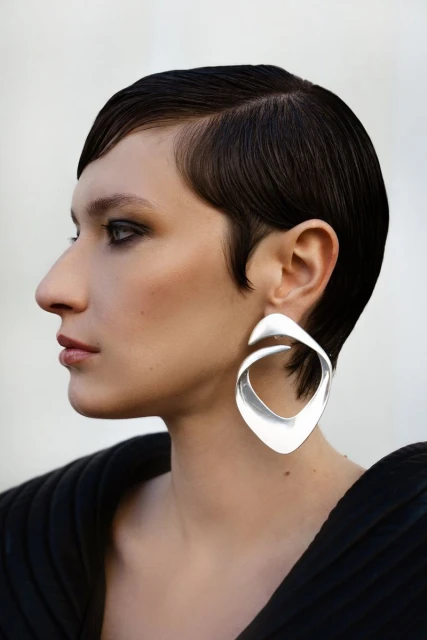 Profile of a woman with a short dark hairstyle wearing a large silver earring and black top against a neutral background.