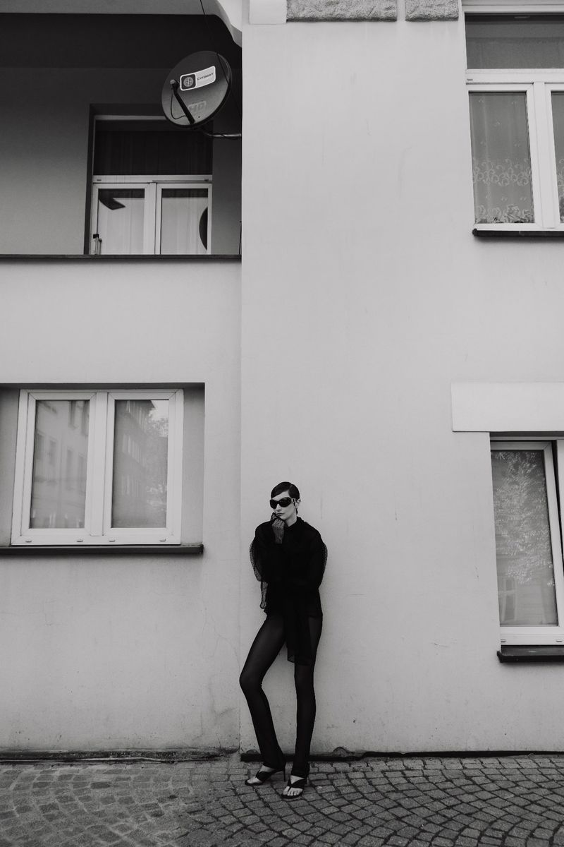 Black and white outdoor portrait of a young adult woman in sunglasses leaning against a white building with a satellite dish.