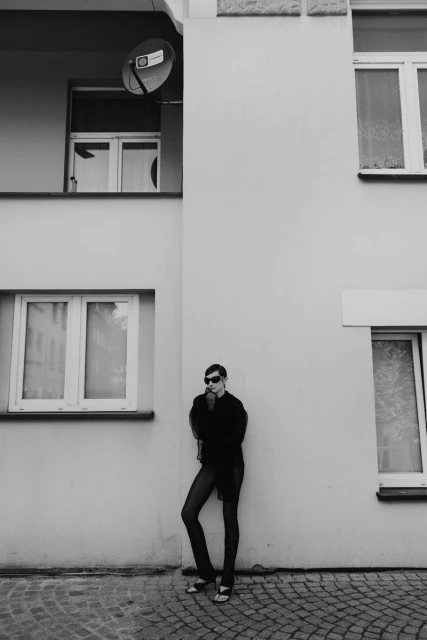 Black and white outdoor portrait of a young adult woman in sunglasses leaning against a white building with a satellite dish.