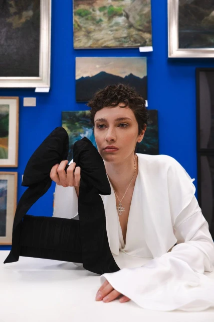 Portrait of woman in white long-sleeved shirt seated at a table holding an oversized black bow with blue wall art background.
