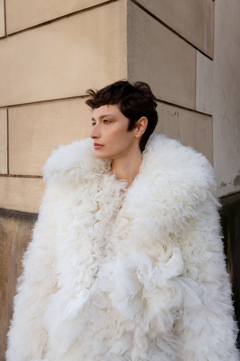 Woman in stylish white ruffled coat poses outdoors against a textured stone wall in soft light, showcasing fashion and style.