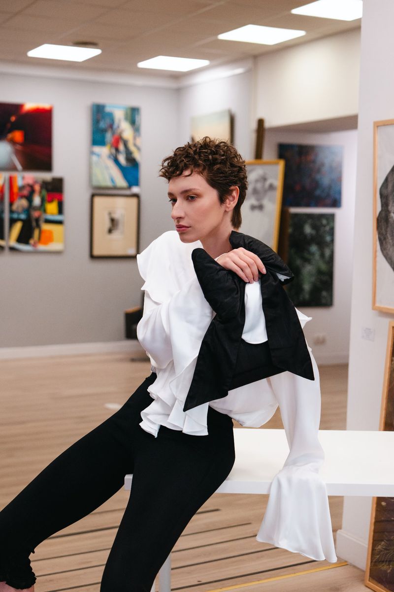 Brunette woman with curly hair poses in a white blouse and black pants, seated on a white surface in a room with artworks on the wall.