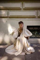 Model wearing a voluminous beige gown with puffy sleeves crouched against a light building exterior wall