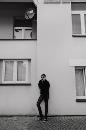A model in an all-black outfit and dark sunglasses leans against the exterior wall of an urban building on cobblestone pavement.