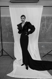 Model in a black structured gown with large shoulder pads posing against a hanging white fabric backdrop outdoors.