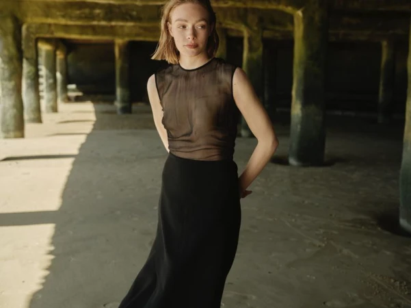 Woman poses wearing a black skirt and sheer top in an underpass. Concrete pillars line the background with light and shadows.