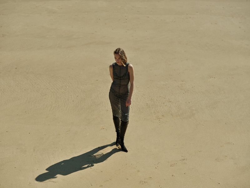 Woman in sheer dress and boots stands on a sandy beach in full sunlight, casting a long shadow in a fashion or editorial shot.