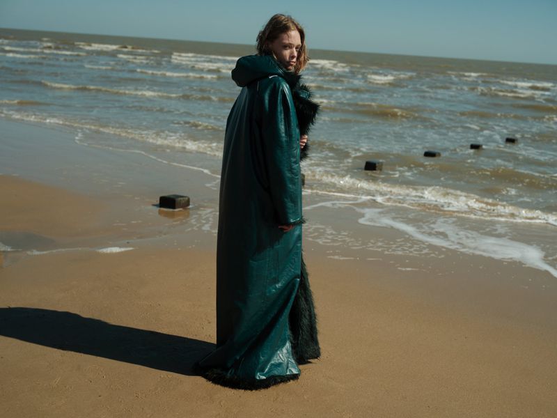 A young adult woman poses on a sandy beach wearing a long, dark green coat with fur trim, with the ocean in the background.