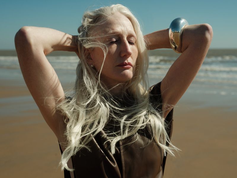 Woman with silver hair poses with arms behind her head on a sandy beach with blue ocean and sky in the background on a sunny day.