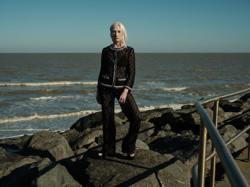 Woman in black lace suit poses on rocks near the ocean under a clear sky; fashion editorial photo with natural light and coastal backdrop.