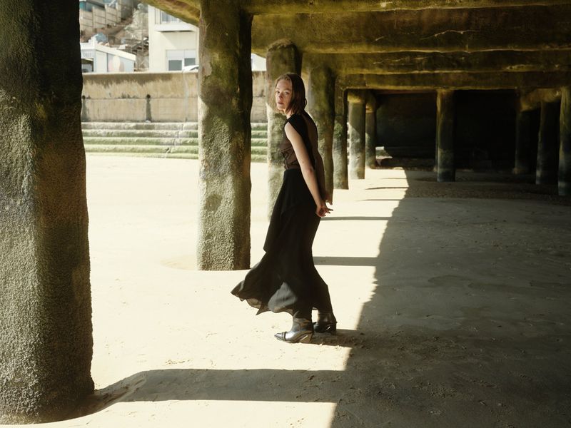 Young woman in flowing black pants and brown top stands under a concrete pier on a sandy beach with visible buildings.