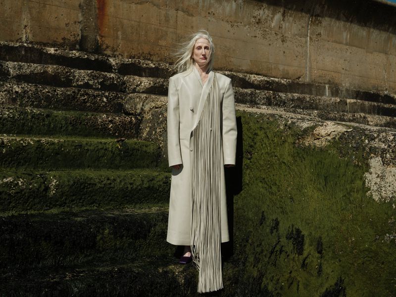 A woman with long gray hair stands on moss-covered concrete stairs in a long, light-beige coat with long fringe detail at the coast.