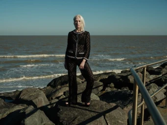 An older woman in a black knit two-piece outfit standing on coastal rocks by the sea under a clear sky.