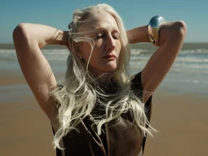 Elderly woman with long white hair wearing a dark brown top and silver cuff on a sandy beach