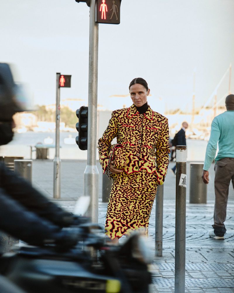 Model Tasha Tilberg wearing a yellow and red patterned skirt suit standing beside a pedestrian signal pole on an urban street.