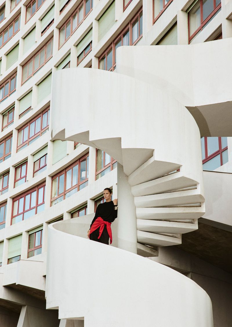 Model Tasha Tilberg in a black outfit with a red sweater tied around her waist standing on a white spiral staircase outside a modernist building.