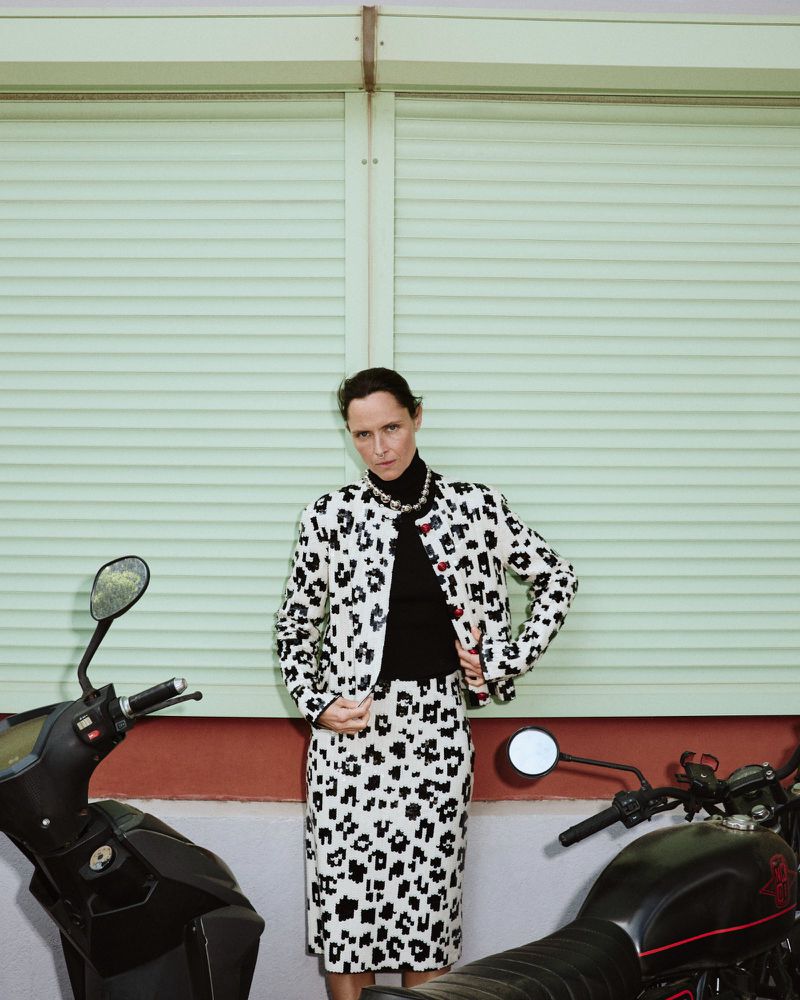 Model Tasha Tilberg poses in a black and white patterned skirt suit and black turtleneck between two parked motorcycles.