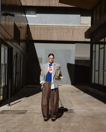 Model Tasha Tilberg wears a plaid blazer and brown wide-leg trousers while posing outdoors in a sunlit architectural alleyway.