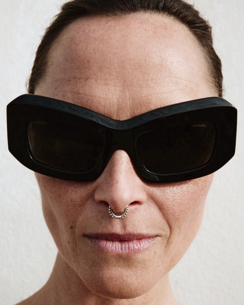 Close-up portrait of model Tasha Tilberg wearing oversized black angular sunglasses and a silver septum ring against a pale backdrop.