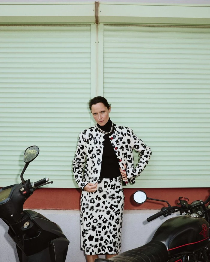 Model Tasha Tilberg poses in a black and white patterned skirt suit and black turtleneck between two parked motorcycles.