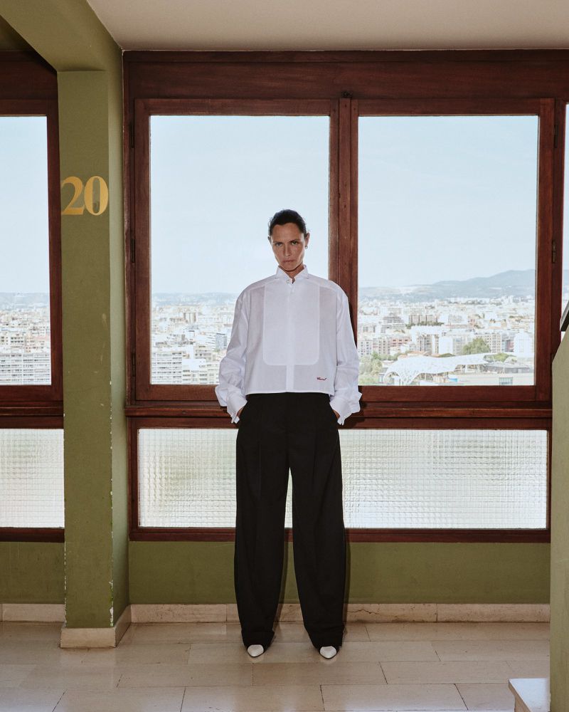 Tasha Tilberg wearing a white cropped bib shirt and wide-leg black trousers standing before large windows overlooking a city.
