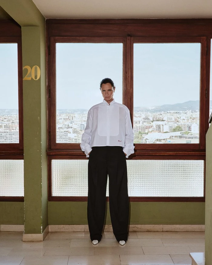 Tasha Tilberg wearing a white cropped bib shirt and wide-leg black trousers standing before large windows overlooking a city.