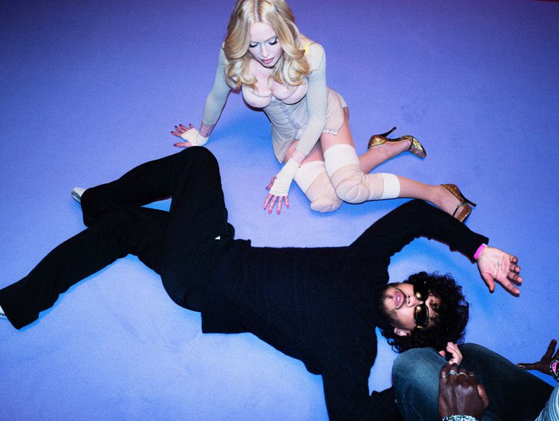 Blonde woman in nude corset and stockings kneeling next to a man lying on a bright blue floor for Vogue Italia
