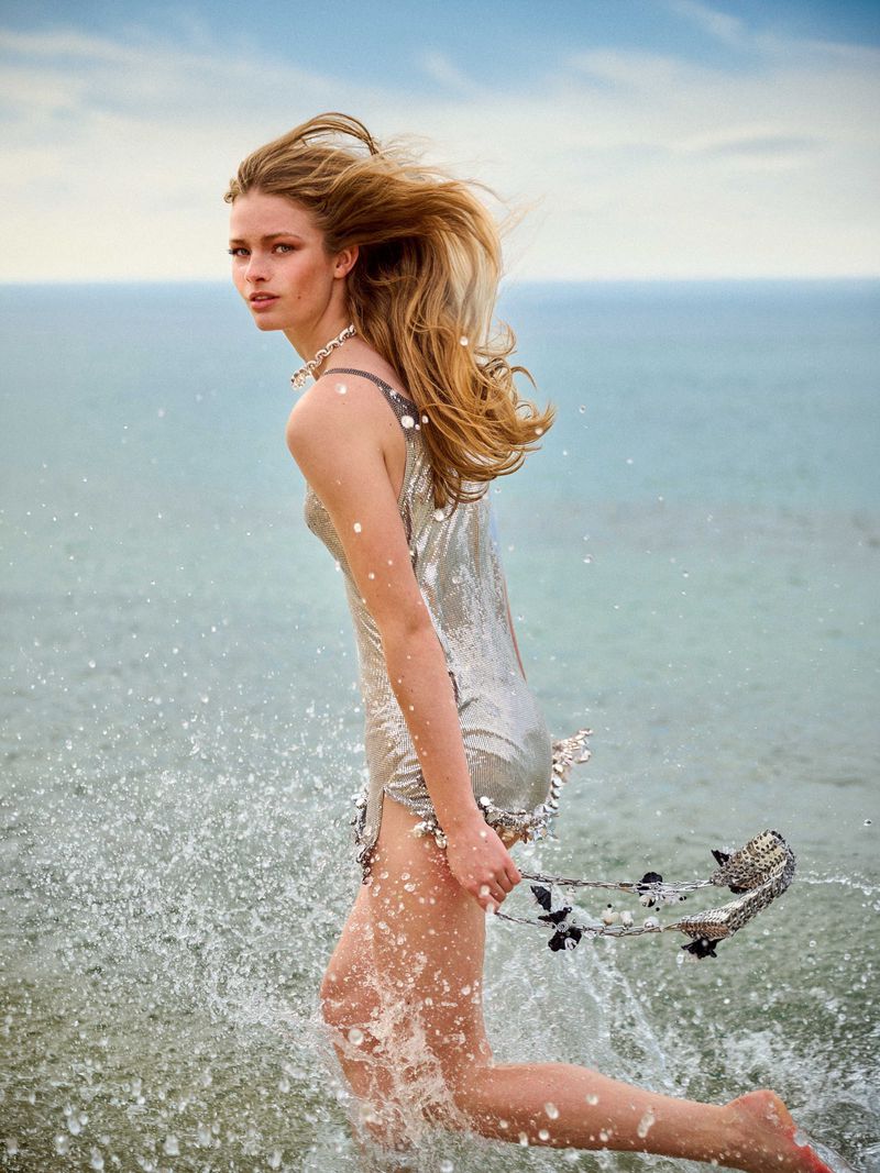 A blonde female model wearing a metallic dress running through ocean water with splashing water droplets around her.