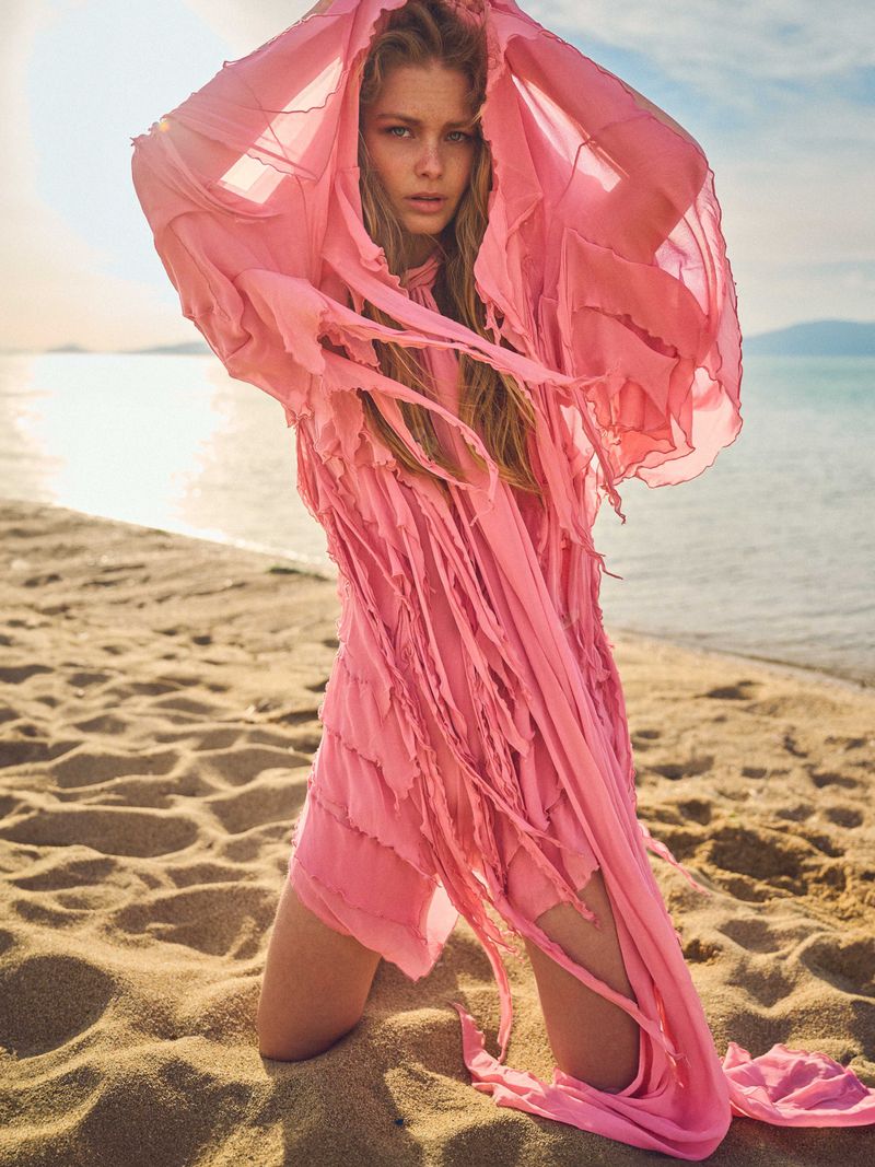 Kate Kraaijeveld kneeling on a sandy beach wearing a sheer pink textured fringed dress with her arms raised.