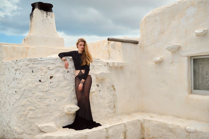 Model Kate Kraaijeveld poses in a sheer black cutout gown on a whitewashed stone rooftop under a cloudy sky.