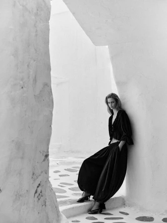 Model wearing dark clothing leaning against a white architectural wall in a narrow sunlit alleyway.