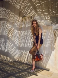 Model Kate Kraaijeveld posing against a sunlit stucco wall with slatted shadow patterns, holding a net bag filled with oranges.