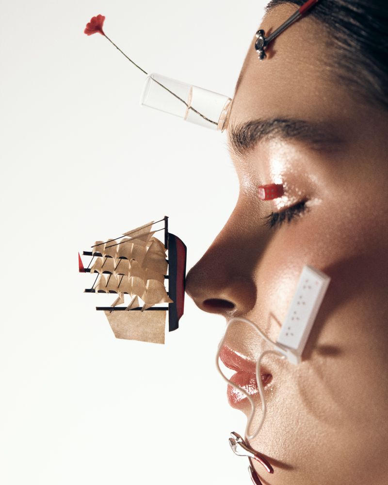 Close-up profile of a woman's face adorned with miniature objects including a tiny sailing ship, power strip, and flower.