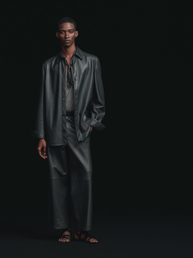 Full-length studio portrait of a male model wearing a dark charcoal leather shirt and matching wide-leg trousers on a black background.