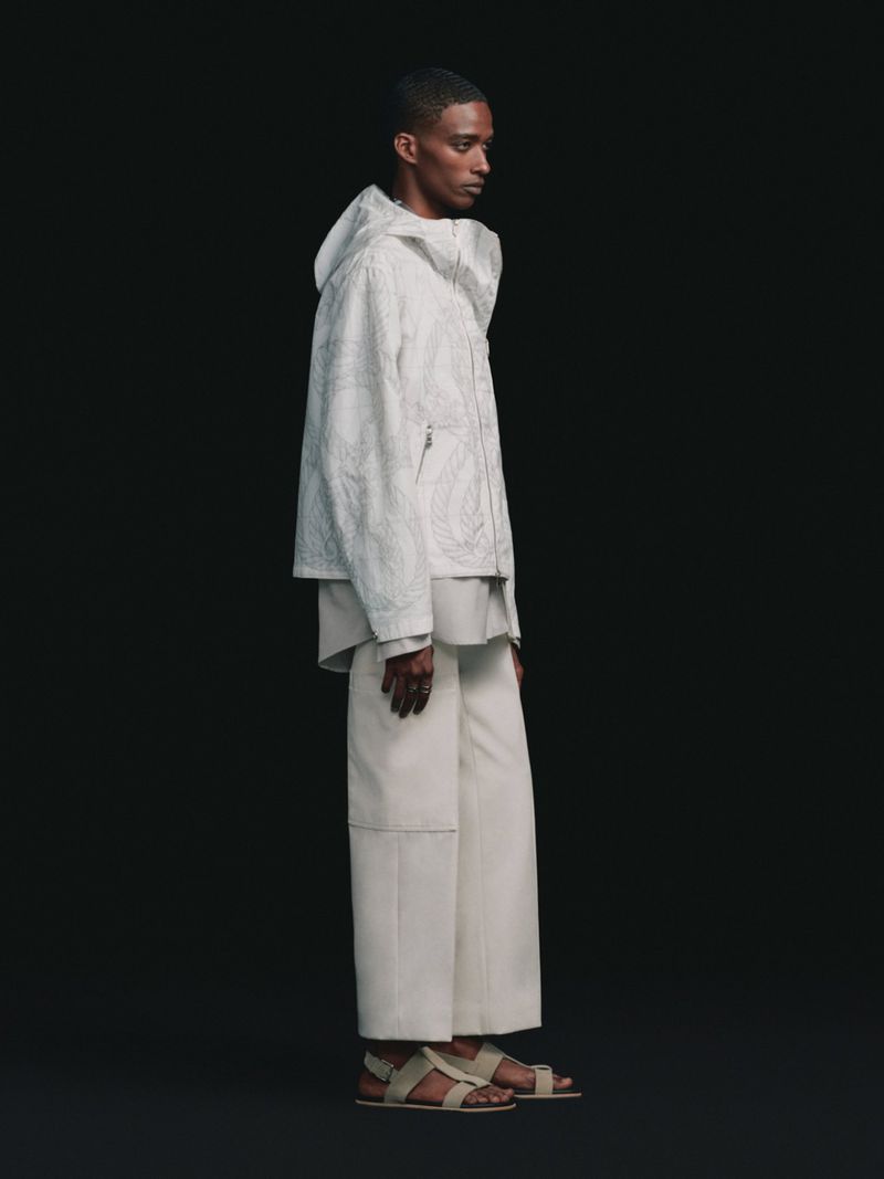 A male model in profile wearing a white hooded jacket, relaxed trousers, and strappy sandals against a solid black background.