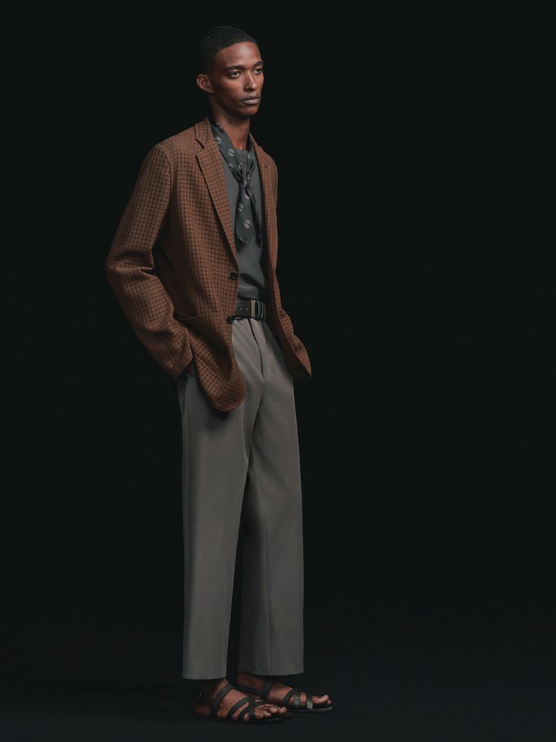 A male model standing in a brown textured blazer, grey tailored trousers, dark neck scarf, and leather sandals against a black backdrop.