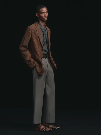 A male model standing in a brown textured blazer, grey tailored trousers, dark neck scarf, and leather sandals against a black backdrop.