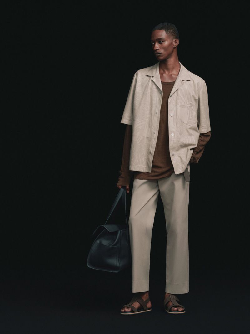 Model posing in a beige short-sleeve shirt, matching trousers, brown undershirt, and leather sandals while holding a dark bag against a black background.