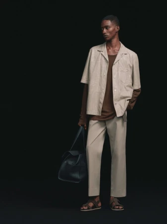 Model posing in a beige short-sleeve shirt, matching trousers, brown undershirt, and leather sandals while holding a dark bag against a black background.