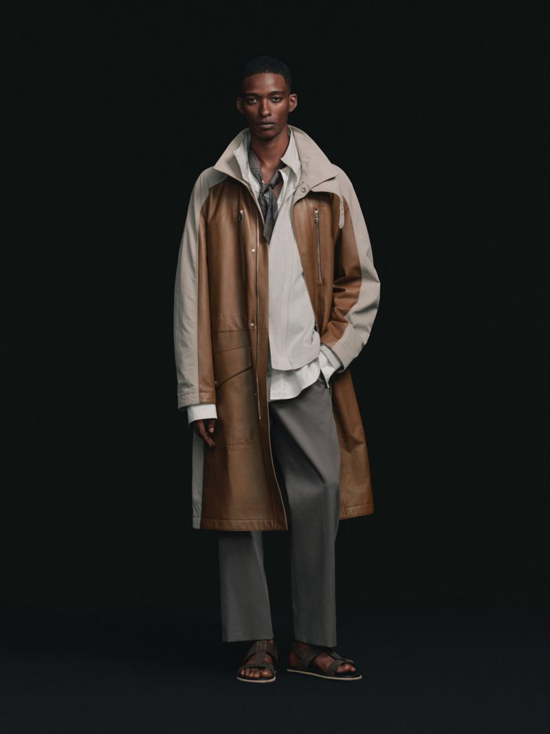 Full-length studio portrait of a Black male model wearing a brown two-tone leather coat, grey trousers, and sandals on a black backdrop.