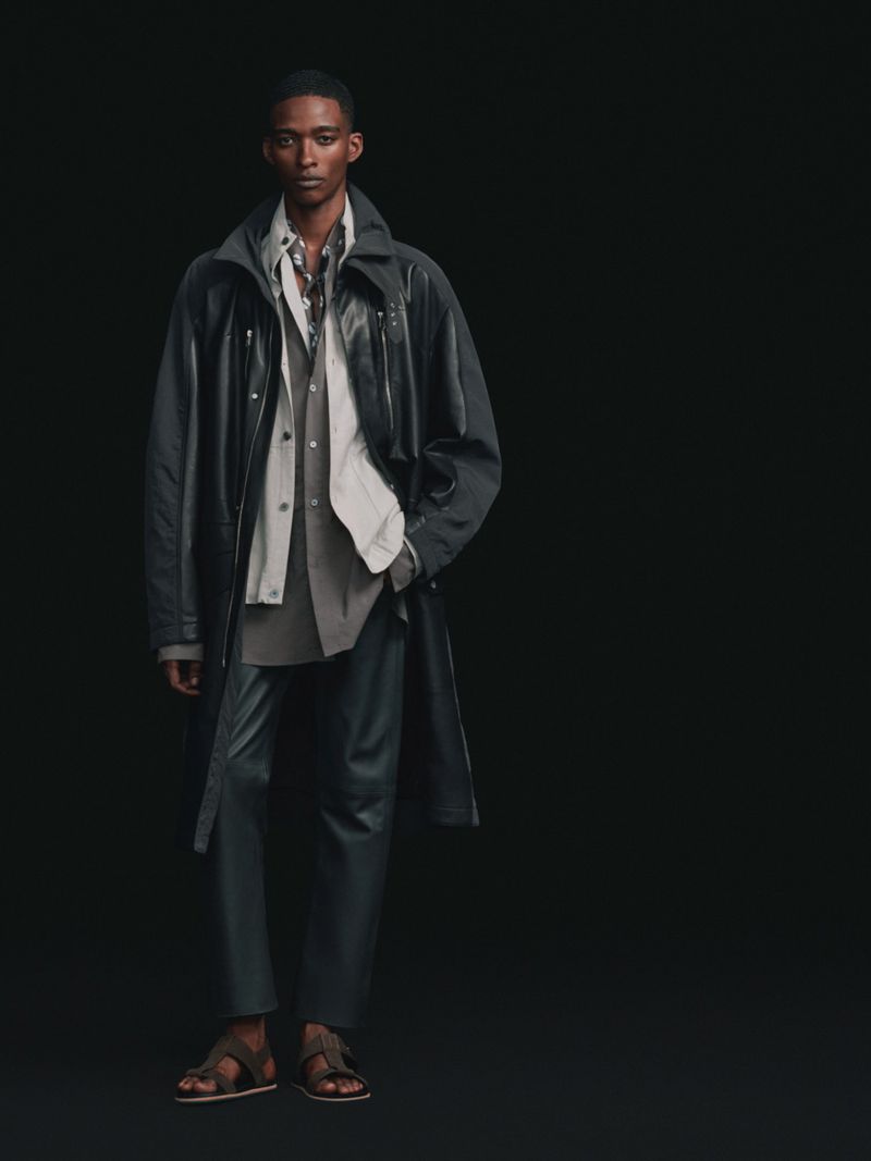 Black male model wearing a dark hooded overcoat, layered shirts, trousers, and sandals against a solid black studio backdrop.