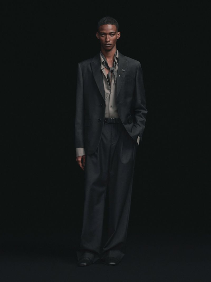 Model wearing a dark tailored suit with wide-leg trousers and a collared shirt against a solid black background.