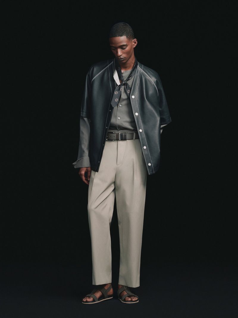 Model posing in a dark short-sleeve leather overshirt, pleated beige trousers, and flat sandals against a black background.