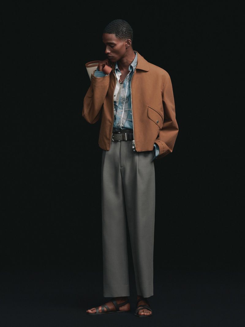 Model standing against a black background wearing a brown jacket, grey pleated trousers, and dark leather sandals with a shoulder bag.