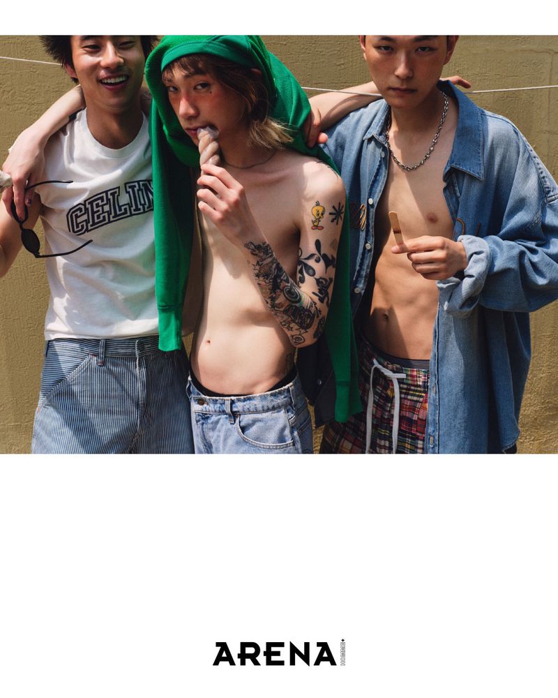 Three young adults in jeans and casual tops pose in an outdoor setting, with one holding a treat and tattoos visible on one arm.