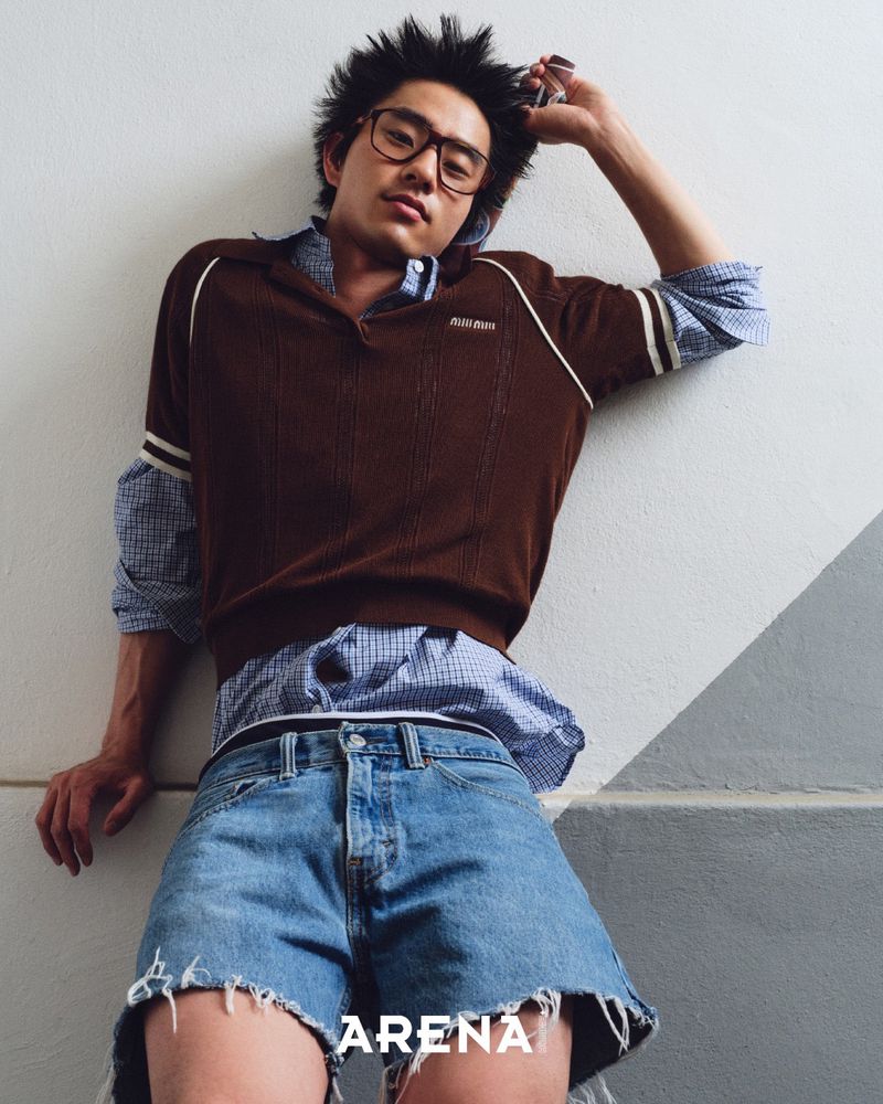 A fashionable Asian male model in a brown sweater and denim shorts reclines against a neutral wall for an indoor editorial fashion shot.