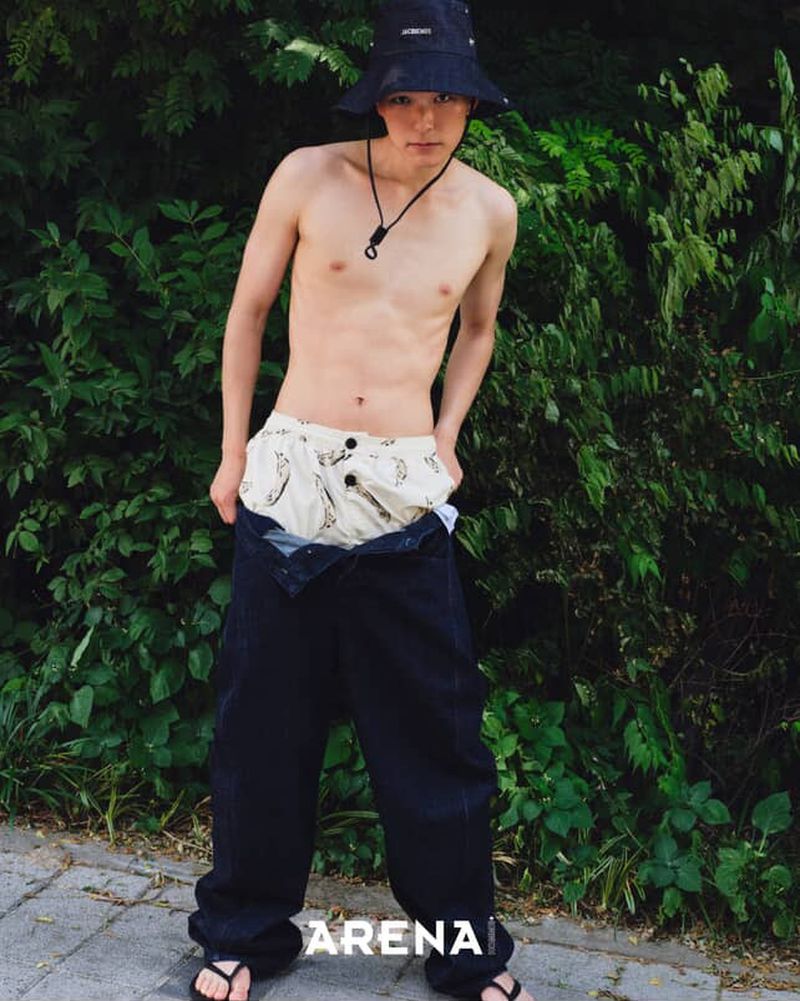 Shirtless young man in bucket hat and sandals poses outdoors with unbuttoned jeans showing patterned boxer shorts against green foliage.